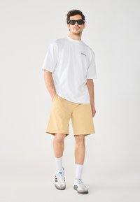 Hvit t-skjorte med korte ermer og svart logo, beige shorts, hvite sokker og sneakers med svarte striper. Modellen har på seg mørke solbriller.