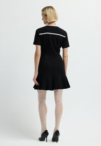 Abito nero con gonna a campana, maniche corte e rifiniture bianche sul retro. Indossato con collant a fantasia e tacchi alti neri.