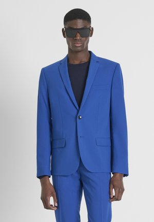 Chaqueta de traje azul a medida con un solo botón, solapas de muesca, dos bolsillos frontales, combinada con una camisa azul marino y elegantes gafas de sol negras.