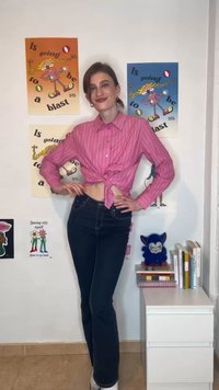 Jonge vrouw in een roze gestreept shirt en donkere jeans poseert glimlachend voor kleurrijke posters en een witte plank met boeken en een blauwe pluche speelgoed.