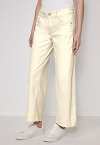 Pantalons en denim larges de couleur crème, avec des poches avant, une fermeture à bouton en métal et une finition lisse et texturée. Associés à des baskets blanches.
