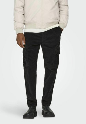 Pantalones cargo de pana negra con bolsillos laterales, corte ajustado y patrón texturizado. Combinados con una chaqueta bomber clara y botas negras.