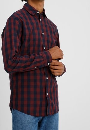 Man wearing a long-sleeve red and black checkered button-up shirt adjusting one cuff, paired with blue jeans against a plain light background.