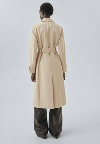 Beige trench coat with a belt, long sleeves, and a vented back, paired with dark brown wide-leg pants. Smooth texture with a tailored fit.