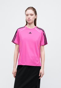 Žena v jasne ružovom športovom tričku Adidas s čiernymi pruhmi na rukávoch a čiernej sukni, stojí na bielom pozadí.