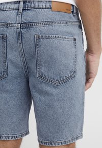 Hátsó nézetben lévő személy világoskék farmer rövidnadrágot visel, hátul zsebekkel és a derékrészén egy világosbarna bőrpamuttal.