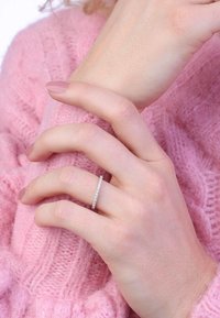 Silberner Ring mit kleinen klaren Steinen, auf einer Hand, die auf einem rosa gestrickten Pullover ruht. Die weiche Textur des Pullovers ist sichtbar.