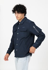 Navyblauwe corduroy jas met een ritszak op de mouw, twee voeringzakken voor en een knoopsluiting. Gelaagd stof en rechte snit.