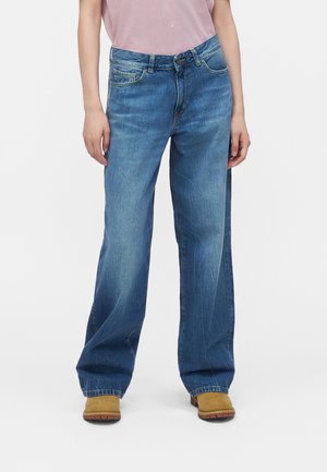 Person iført løstsiddende blå jeans og brune støvler, stående mod en hvid baggrund, hænder afslappet langs siderne.