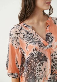 Blouse à manches courtes et décolleté en V en corail clair, ornée de motifs paisley noir et blanc complexes. Tissu léger avec une texture douce.