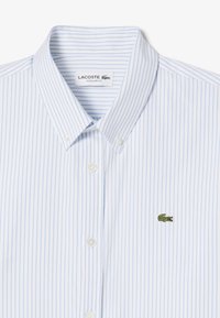 Camisa de rayas blanca y azul claro, con cuello y botón en la parte frontal, que presenta un pequeño logo de cocodrilo verde en el lado izquierdo del pecho.