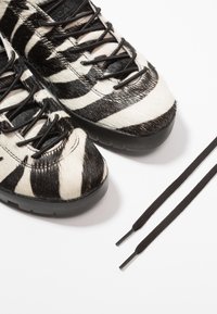 Zapatillas con textura de piel a rayas blanco y negro, suelas y cordones de goma negra, con detalles lisos y diseño de punta redondeada.