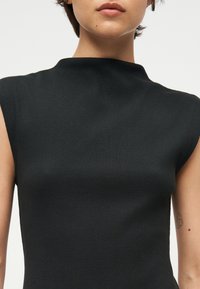 Gros plan d'une personne portant un haut noir côtelé sans manches avec un col montant et un petit tatouage sur le bras droit supérieur.