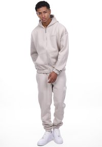 Beige zip-up hættetrøje med hætte og forlommer, sammen med matchende tapered sweatpants. Har broderet logo og ribbede manchetter.