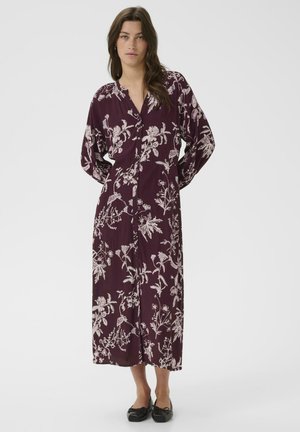 A woman with long hair stands wearing a long, burgundy dress adorned with white floral patterns and black shoes, posing against a plain background.