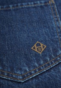 Mörkblå denimtyg med en texturerad yta, med en liten, beige, broderad logotyp i diamantform nära en fick söm.