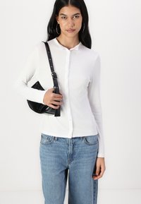 Chemise blanche à manches longues côtelées avec fermeture à boutons, associée à un jean bleu clair ample. Tenant un petit sac à main noir avec une bandoulière.