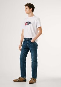 Camiseta blanca de manga corta con un estampado de logo colorido, combinada con jeans de mezclilla azules y zapatillas marrones con acentos en azul y blanco.