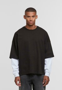 Sort oversized t-shirt med korte ærmer, lagt over en hvid langærmet skjorte. Blødt stof, rund halsudskæring, afslappet pasform.