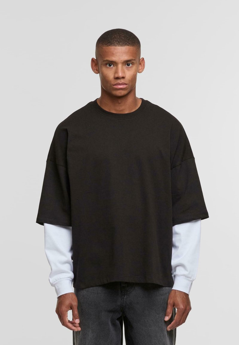 Sort oversized t-shirt med korte ærmer, lagt over en hvid langærmet skjorte. Blødt stof, rund halsudskæring, afslappet pasform.