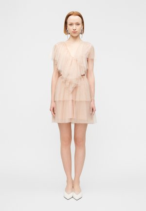 Mini sukienka z falbanami z tiulu w delikatnym brzoskwiniowym kolorze, z warstwowymi falbanami, głębokim dekoltem w serek i przezroczystymi rękawami. Do tego białe szpiczaste szpilki.