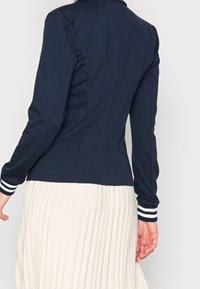 Granatowy top z długim rękawem o dopasowanym kroju, z małym kołnierzykiem i białymi paskami na mankietach. Gładka faktura materiału.