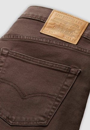 Jeans de mezclilla marrones con una etiqueta de cuero que presenta texto en relieve, bolsillo trasero con un patrón de costura distintivo y textura suave.