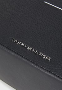 Portafoglio in pelle nera con texture, caratterizzato dal logo "TOMMY HILFIGER" in rilievo argento e dettagli a striscia orizzontale bianca.