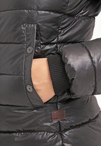 Ruka v kapse černé prošívané bundy se zapínáním na druky a žebrovanými manžetami, s hnědou nášivkou G-Star Originals na přední straně.