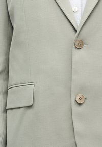 Κοντινό πλάνο ανοιχτοπράσινου σακακιού με δύο καφέ κουμπιά, ένα τσεπάκι με καπάκι και μέρος ενός λευκού πουκαμίσου από κάτω.