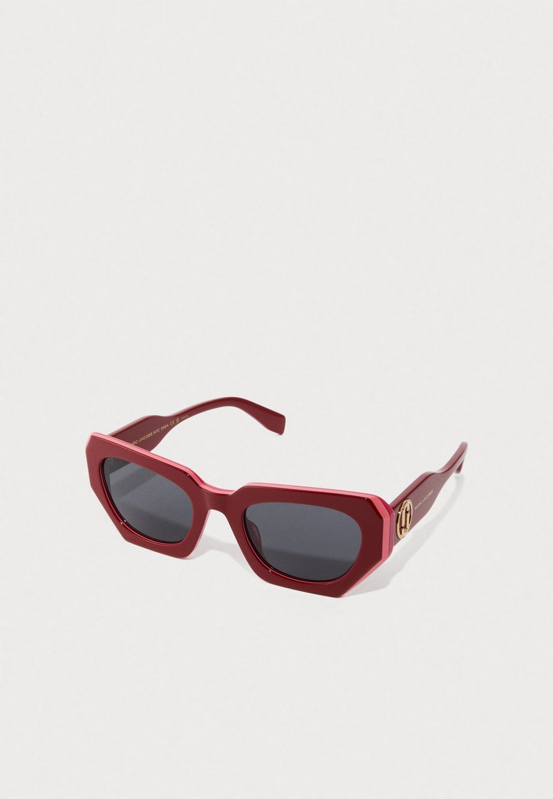 Red oversized hexagonal sunglasses with dark lenses and gold logo detail on the temples, resting on a white surface.