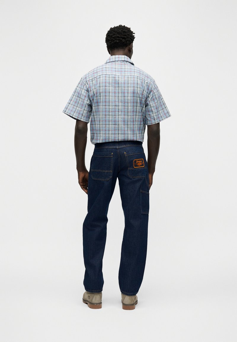 Mann von hinten stehend, trägt hellblaues kurzärmeliges kariertes Hemd, dunkelblaue Jeans mit Gesäßtaschen und beige Schuhe.