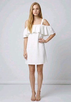 Mujer joven de pie con la mano en la cadera, vestida con un vestido blanco con hombros descubiertos y volantes, y tacones abiertos beige sobre un fondo liso.