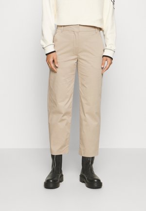Personne portant un pantalon beige à jambes droites et des bottines noires, debout devant un fond clair uni.