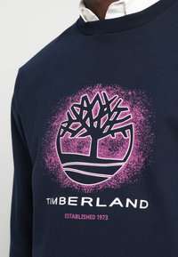 Hanorac bleumarin cu un logo stilizat de copac alb și roz și textul "Timberland Established 1973" deasupra unui tricou alb cu guler.