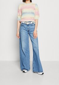 Wielokolorowy sweter dziany z krótkimi rękawami, z pastelowymi paskami i ażurowym wzorem. Połączony z długimi jasnoniebieskimi dżinsami i białym paskiem.