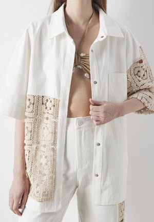Kvinde iført hvid oversized skjorte med hæklet lapper, matchende højtaljede bukser og en guldfarvet halter bikini top med perledetaljer.