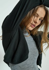 Černý oversized kardigan s dlouhými rukávy, vyrobený z měkké pleteniny. Nosený přes světle šedé těsné triko, které vykazuje hladkou texturu a ležérní design.