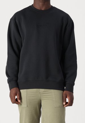 Homme portant un sweatshirt noir à col ras du cou avec un logo subtil en relief et un pantalon vert clair avec des poches avant.