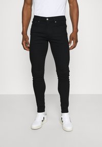 Vaqueros ajustados negros de mezclilla, con un clásico diseño de 5 bolsillos, cierre de botón metálico y una textura suave. Combinados con zapatillas blancas.