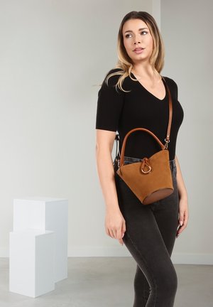 Vrouw gekleed in een zwart topje en spijkerbroek, met een bruine suède bucket bag met leren handvat en riem, staand voor een grijze achtergrond.
