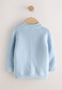 Pull en maille côtelée bleu clair à manches longues accroché sur un cintre en bois contre un mur blanc.