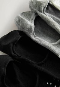 Trois paires de chaussettes invisibles en noir, gris et blanc, superposées sur une surface claire.