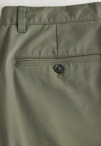 Tissu vert olive avec passants de ceinture cousus et détail de poche arrière boutonnée sur un pantalon.