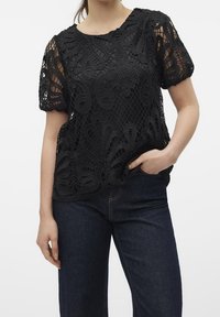 Haut en dentelle noire avec un motif floral, manches courtes, encolure ronde et coupe ample, associé à un jean en denim foncé.