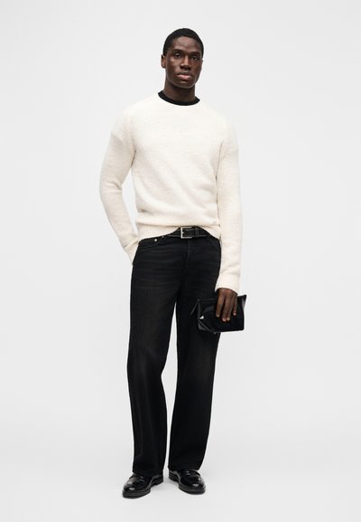 White textured sweater, black jeans, black belt, and black shoes. Model holds a black clutch. Simple, modern, and monochromatic look.