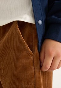 Hand aan de zijkant rustend, gekleed in een paarse corduroy broek, een wit ribbed shirt en een marineblauwe cardigan met knopen.