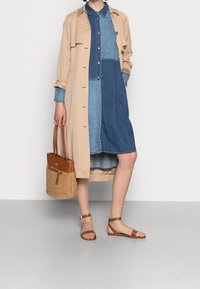 Beżowy trencz nałożony na sukienkę koszulową z denimu w niebieskie i jasnoniebieskie panele, połączony z brązowymi sandałami i plecioną torebką w odcieniu jasnobrązowym.