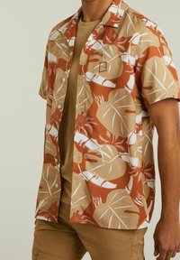 Camicia a maniche corte marrone e beige, con un motivo di foglie tropicali bianche; tessuto leggero e vestibilità rilassata.