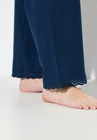 Námořnické modré bavlněné kalhoty s vlnitým krajkovým lemem na spodním okraji, vykazující hladkou texturu a široké nohavice. Viditelné bosé nohy.